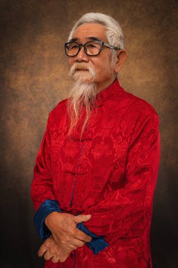 Stüdyo arka planında geleneksel Çin kıyafetleri içinde beyaz saçlı ve sakallı gözlüklü Çinli ihtiyarın stüdyo portresi.