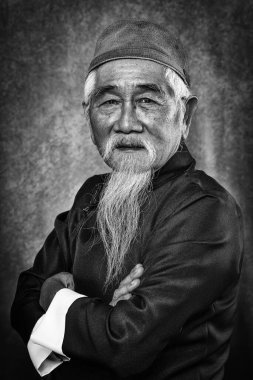 Stüdyoda, beyaz saçlı ve sakallı, geleneksel Çin kıyafetleri içinde, siyah beyaz arka planda Çinli yaşlı bir adamın portresi.