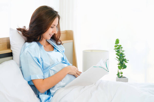 portrait of happy pregnant woman sitting on bed reading book at home in bedroom