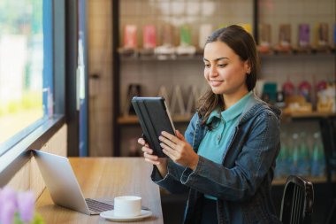 Kahve dükkanının vitrininde oturan güzel hipster portresi elinde tablet, elinde kahve fincanı ve bilgisayarla tezgahta duruyordu.