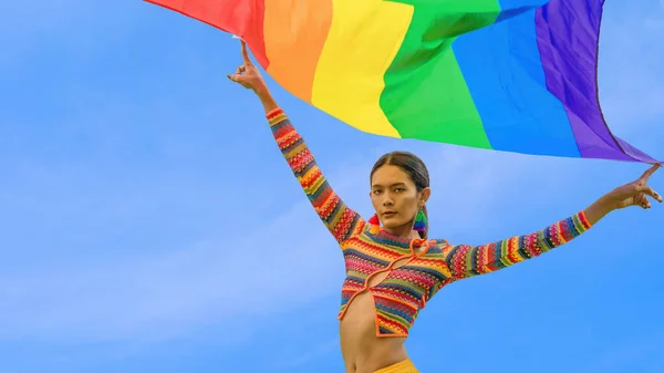 Portrait of transgender person posing with pride flag with blue sky background in celebration of pride month for human equality and diversity for lgbtq people