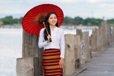 portrait of Myanmar woman in traditional dress holding umbrella standing at U Bein bridge at Amarapura Manndalay