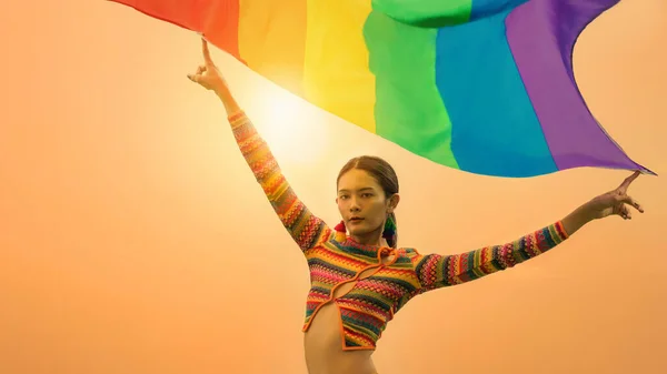 portrait of asian transgender person in colorful dress posing with rainbow pride flag to celebrate of LGBTQIA right in equality and diversity in pride month with background of sunrise sky