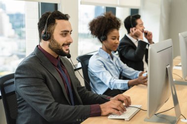 Ortadoğu erkek çağrı merkezinin portresi ya da çağrı merkezinde çalışan yardım masası ya da telefon destek ajanı. Kulaklıkla diğer ajanların bulanık arka planlarını takıyor.