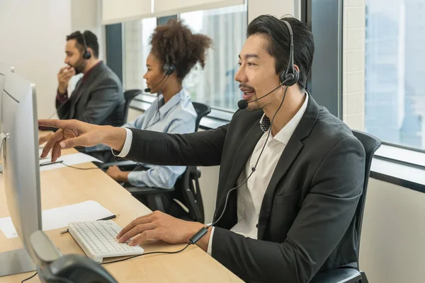 portrait of asian male call center or help desk or telephone support agent working at call center workplace wearing headset with blurred background of other agents working