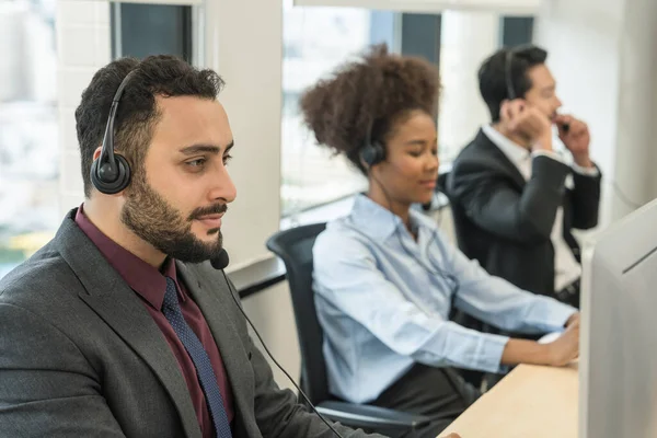portrait of middle east male call center or help desk or telephone support agent working at call center workplace wearing headset with blurred background of other agents working