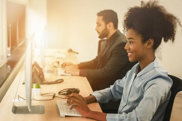 portrait of smiling mixed race female call center or help desk or telephone support agent working at call center workplace wearing headset with blurred background of male agent working with light flare