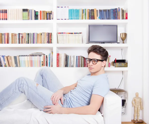 Sexy hombre con gafas acostado en el sofá en frente de estante de libros —  Fotos de Stock