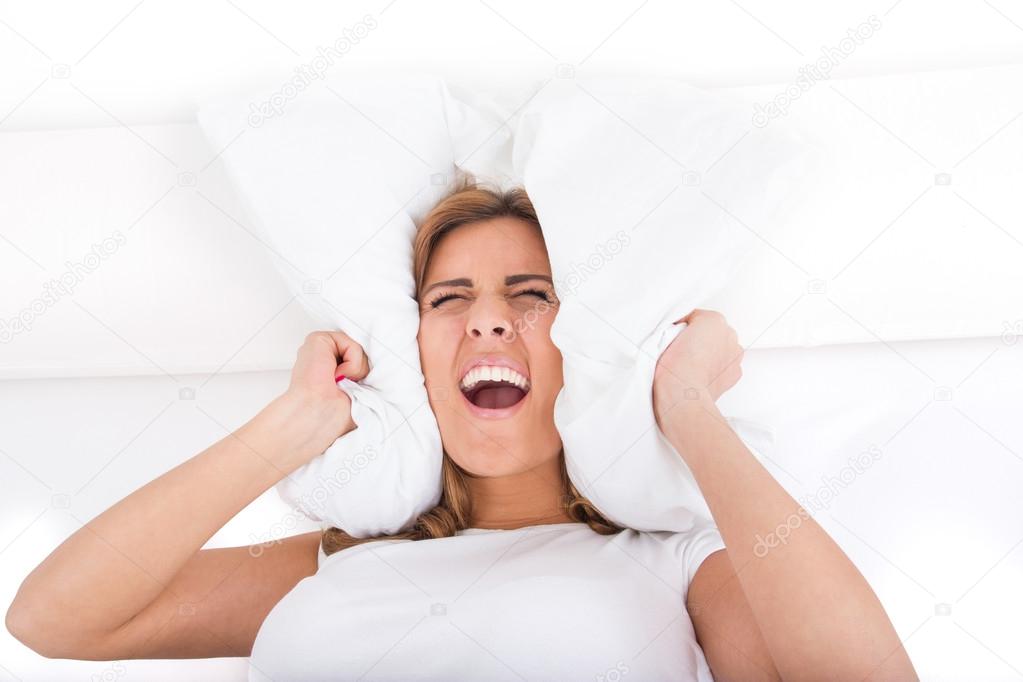 Woman screaming and covering her ears with pillow because of noi