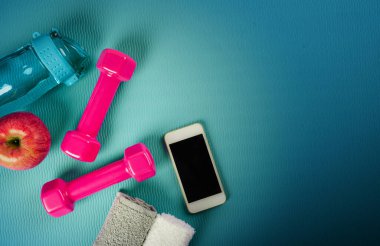 Spor aletleri. Pembe dambıl, havlu, taze elma, akıllı telefon ve mavi turkuaz yoga minderinde bir şişe su. Eğitim, egzersiz ve spor konsepti. Fotokopi alanı ile mavi arkaplanda düz resim yatıyordu.
