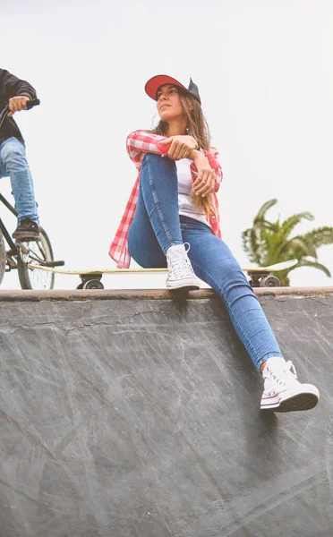 Mooie jonge vrouw in cap zit op skatepark op de helling met haar longboard. — Stockfoto