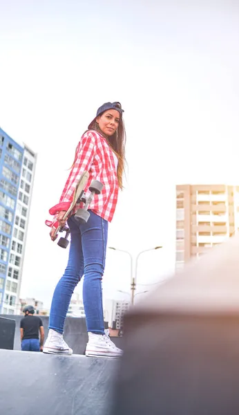 Bella giovane donna in berretto è seduto a skatepark sulla rampa con il suo longboard. — Foto Stock