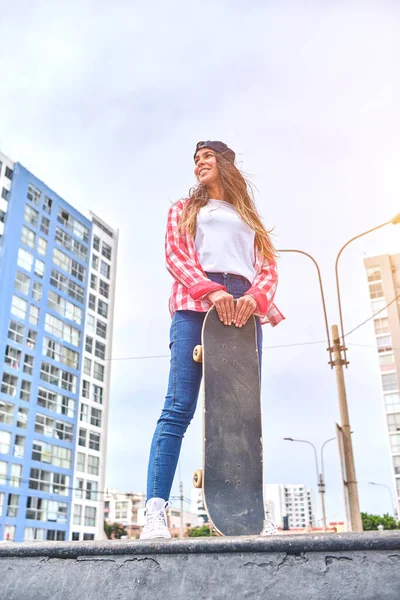 Mooie jonge vrouw in cap zit op skatepark op de helling met haar longboard. — Stockfoto