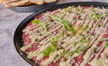 Carpaccio bifteği, parmesanlı soğuk aperatif, kapari ve roka, siyah tabakta seçici odaklanma.