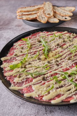 Carpaccio bifteği, parmesanlı soğuk aperatif, kapari ve roka, siyah tabakta seçici odaklanma.