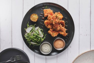 Kızarmış tavuk, gurme roka ve turp salatası, yanında da Peru biber sosu, siyah bir tabakta servis ediliyor..