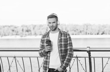 Kareli tişörtlü ve modern kulaklıklı genç yakışıklı açık havada müzik dinlerken kahve içiyor.