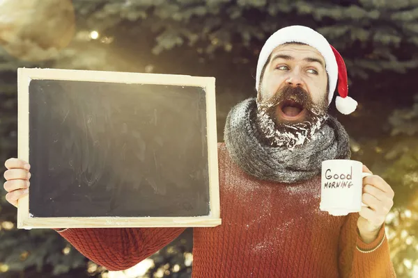Knappe gelukkige man in Santa Claus hoed, bebaarde hipster met baard en snor bedekt met witte vorst, houdt blank zwart bord en koffie kopje winter dag buitenshuis op natuurlijke achtergrond, copy Spa — Stockfoto