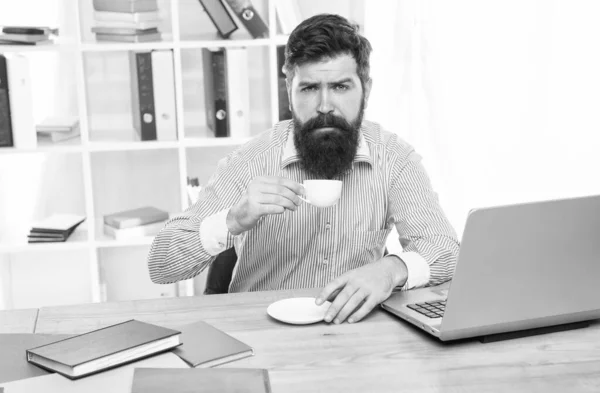 Uitgeputte kantoorbediende met hippe baard drinkt koffie op het bureaublad in modern kantoor, 's morgens — Stockfoto