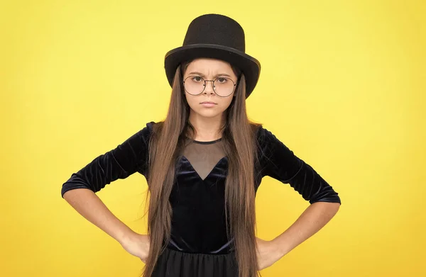 Serious female baby in glasses with long hair wear fashion party dress and hat standing with hands on hips yellow backgroud, girl — Stock Photo, Image