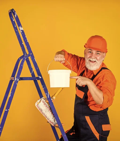 Boyalı kovayla şapkalı bir ressam. İnşaat işçisi boyama cephesi. Isı yalıtımı işe yarıyor. Boyacı merdiveni. Onarım yapmak için. İş elbiseli bir adam boş odada duvar boyuyor.