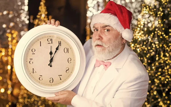 Štědrý večer. Santa Claus počítá čas. Půlnoční koncept. Nový rok odpočítávání. Muž s retro hodinami. Pozadí osvícení věnce. Vousatý obchodník v Santově klobouku. Senior Santa Man ve smokingu — Stock fotografie