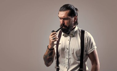 resmi erkek tiryaki. Askılı ve papyonlu eski moda sakallı hippi pipo içiyor. Kendine güvenen ve klasik giyinen zarif bir adam sigara piposu giyer. Erkeklik ve karizma. Boşluğu kopyala