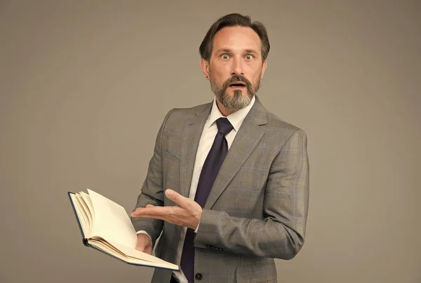 Profesor de escuela senior en traje formal apuntan mano en libro abierto con aspecto sorprendido fondo gris, enseñanza — Foto de Stock