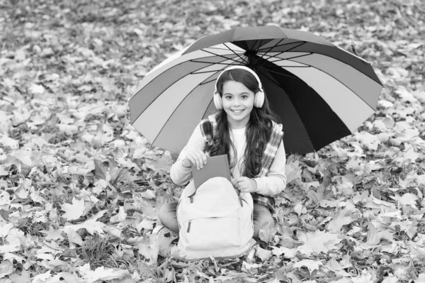 Menina feliz ouvir música no fone de ouvido enquanto sentado na floresta de outono entre folhas caídas com guarda-chuva colorido e mochila escolar vai ler o livro, relaxar — Fotografia de Stock