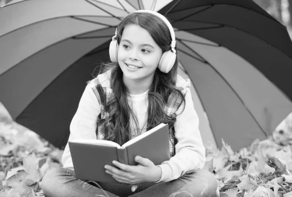 Dia de outono perfeito de menina alegre sob guarda-chuva colorido ouvir música e ler livro em fones de ouvido e sentado no parque temporada de outono desfrutando de bom tempo, literatura infantil — Fotografia de Stock