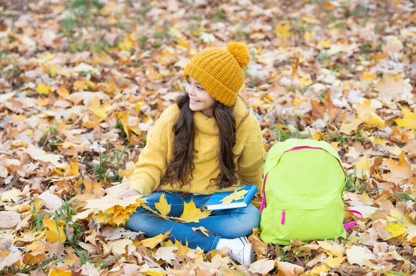 Sorridente ragazza adolescente in cappello giallo tenere foglia d'acero con zaino scuola rilassarsi nel parco, tornare a scuola. — Foto Stock