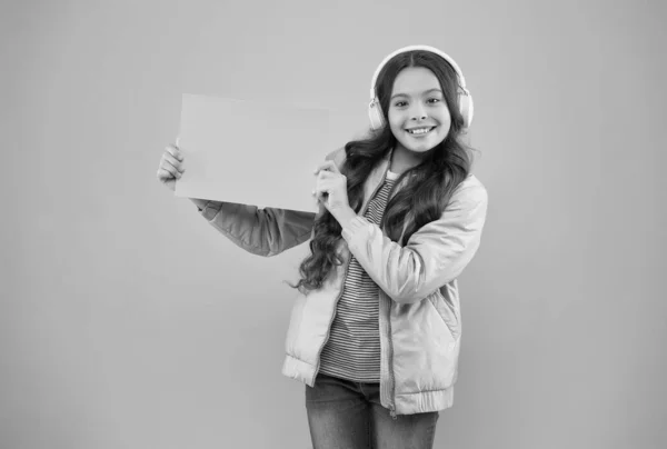 Cheerful kid wearing earphones and showing empty paper for copy space, advertisement — Stock Photo, Image