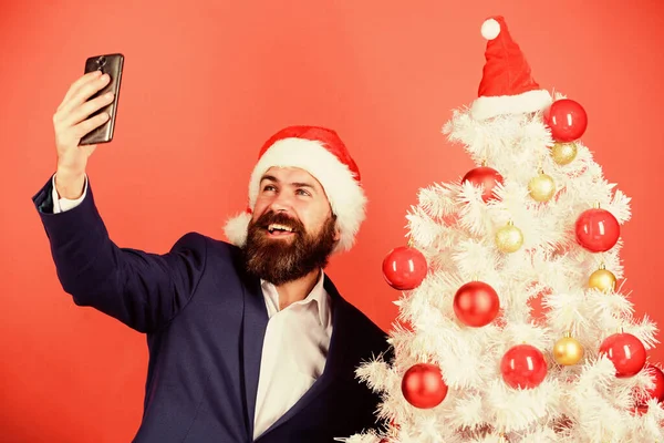 Selfie w pobliżu choinki. Uchwyć szczęśliwą pamięć. Przyjęcie noworoczne. Wesołych Świąt. Brodaty w sieci. biznesmen pracujący na wakacjach. Pozdrawiam jego najbliższych. manager santa hat używać smartfona — Zdjęcie stockowe