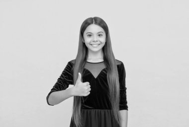 Moda elbiseli, uzun saçlı mutlu küçük çocuk el işareti sarı arka plan, baş parmak işareti