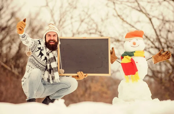 Anunţ de iarnă. Hipster tricotat pălărie și mănuși arată tablă. Sărbători de iarnă. Guy și om de zăpadă fundal natură înzăpezită. Eveniment de iarnă. Eveniment viitor. Omul cu barbă ține creta copia spațiu — Fotografie, imagine de stoc