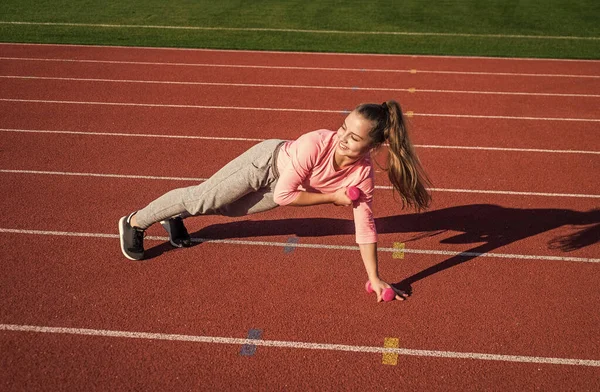 Güç ve denge. sağlıklı çocukluk. Açık havada egzersiz yapmak için. Halterli kendine güvenen bir sporcu. Genç kız stadyumda ısınıyor. Spor giyim bölümündeki çocuk dambıl kullanıyor. Çocuk yarış pistinde egzersiz yapıyor. — Stok fotoğraf