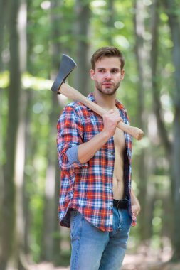 Sert bir görünüm. Tıraşsız bir adam ormanda balta taşır. Oduncu tarzı. Baltalı adam açık ekose gömlek ve kot pantolon giyer. Sıradan bir tarz. Doğa yolculuğu. Yaz modası. Moda tarzı. Her gün gardırop