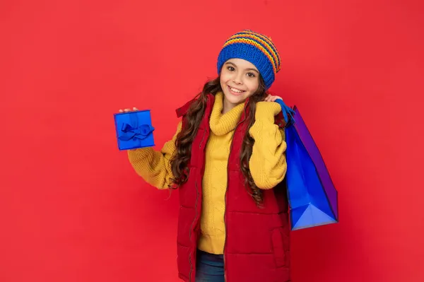 冬の帽子の幸せな子供赤い背景にショッピングバッグやギフトボックスを保持し、購入 — ストック写真