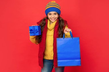 Kış kıyafetleri içinde mutlu çocuk alışveriş çantası ve kırmızı arka planda kutu, satın al