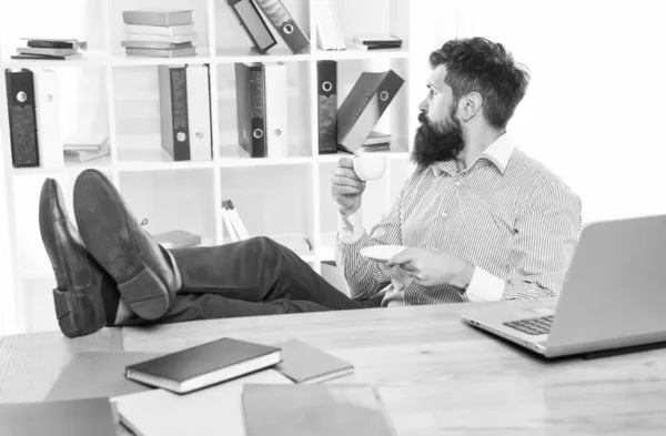 Bärtiger Mann mit brutalem Blick entspannt sich am Schreibtisch und trinkt Kaffee im modernen Büro, Hipster — Stockfoto