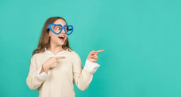 Gelukkig tiener meisje in partij bril wijzen vinger op blauwe achtergrond met kopieerruimte, zomer mode — Stockfoto