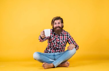 Vahşi sakallı adam kareli gömlek giyiyor, gür sakallı ve bıyıklı sabah kahvesini fincandan içiyor.