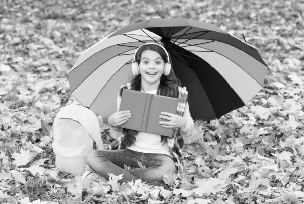 Enfant adolescent joyeux se détendre dans la forêt d'automne ou parc tout en écoutant de la musique dans le casque et lire le livre près du sac à dos de l'école sous parapluie coloré, automne — Photo