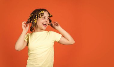 Moda ipuçları. Kıvırcık saçlı, turuncu saçlı küçük bir kız. Sevimli çocuk saçı. Gençlik hobileri. Kafasında bigudileri olan kızı gülüyor. Kuaför salonu. Kadın güzellik rutini