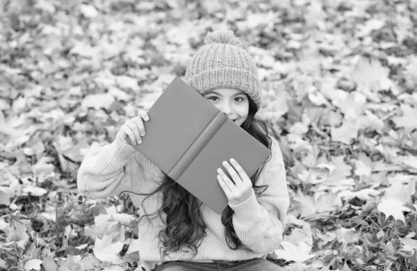 Happy little kid look behind school book sitting on fall leaves outdoors, education — Stok Foto