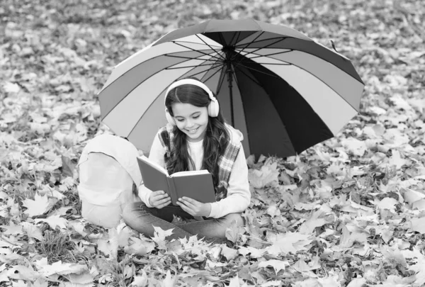 在漫长的一天之后休息。快乐的童年回学校去戴耳机的女孩在公园里休息一下。在森林里享受秋天的乐趣。听音乐。在线课程教育。秋天的孩子在伞下。读这本书 — 图库照片