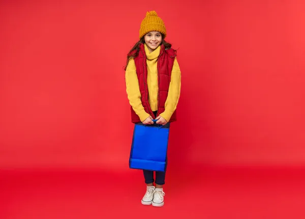 Счастливый ребенок в зимней шляпе держать сумки для покупок на красном фоне, покупки — стоковое фото