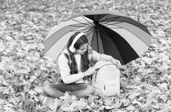 幸せな子供は秋の森の中にカラフルな傘と学校のバックパックで座っている間にイヤフォンで音楽を聴く秋 — ストック写真