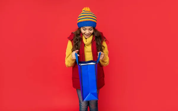 크리스마스에 빨간 배경으로 쇼핑 가방을 열어 놓은 겨울 옷을 입은 아이가 놀라는 모습 — 스톡 사진