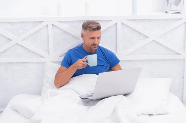 Glad kille koppla av dricka morgon te och använda laptop dator i sängen, koppla av — Stockfoto